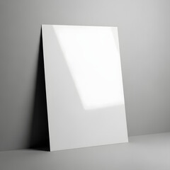 A blank white poster leaning against a gray wall, illuminated by natural light.