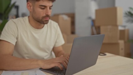 Man typing on laptop in building surrounded by shipping boxes and paperwork spread on table; calm concentration. - Powered by Adobe