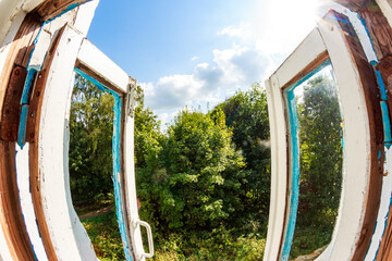 Rustic abandoned window frame, peeling paint, seen from inside, revealing lush summer greenery and a brilliant blue sky with sunlight