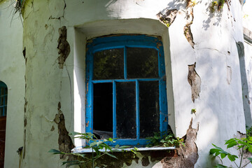 Weathered blue window in a decaying manor wall. Peeling stucco, crumbling facade, and vibrant green foliage signal nature's reclaim. Summer abandonment