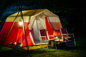 Camping tents at night, vintage cabin tents, antique oil lamps, retro chairs, and a group of camping tents with outdoor coffee-making facilities on wooden tables in a forest camping area.