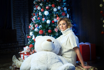 A woman in a white sweater is sitting on the floor with a white teddy bear near a Christmas tree.