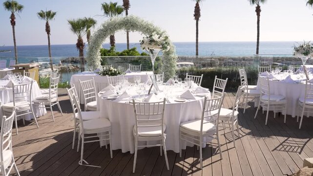 Beautiful outdoor wedding setup with round tables dressed in white linens. A floral arch frames the stunning sea view, creating a perfect atmosphere for a celebration by the shore.