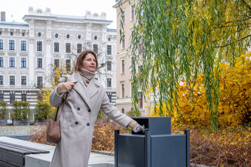 Mature woman in stylish beige coat discarding waste into modern trash bin surrounded by autumn foliage and elegant architecture, promoting environmental awareness and responsibility