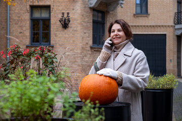 Mature woman in a cozy beige coat, smiling while talking on the phone, stands beside a vibrant pumpkin in an autumn garden with colorful plants and charming architecture