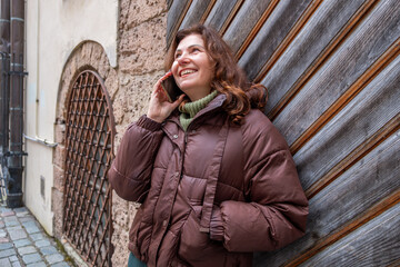 Midlife woman with brown hair, wearing a cozy jacket, smiles while talking on her smartphone, standing against a textured wooden wall, conveying warmth and connection in an urban setting