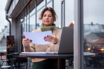 Middle-aged woman engaged in remote work at a food court, reviewing documents while seated at a table with a laptop and a cozy atmosphere, showcasing modern work-life balance