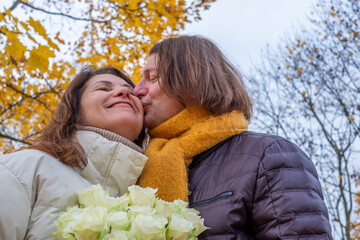 Midlife couple sharing a tender moment in autumn, surrounded by vibrant yellow leaves, holding a bouquet of white roses, celebrating love and connection in a serene outdoor setting