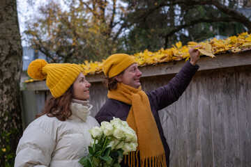 Midlife couple enjoying autumn scenery, wearing matching yellow hats, holding roses, admiring colorful leaves, creating a romantic atmosphere with nature's beauty and seasonal charm