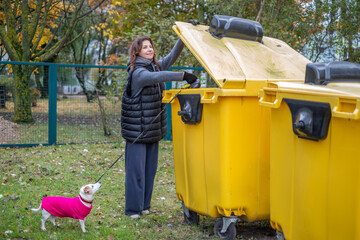 Middle-aged woman in a black vest lifts lid of yellow dumpster while walking her Jack Russell Terrier in a park, showcasing daily life and responsible pet ownership