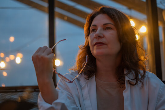 Mature woman with glasses, enjoying a thoughtful moment in a cozy cafe during evening hours, surrounded by warm lighting and a relaxed atmosphere, reflecting on life experiences - Powered by Adobe