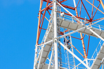 A close-up view of a telecommunication tower with a clear blue sky background. The image showcases the intricate metal framework and ladders used for maintenance. © SerPak