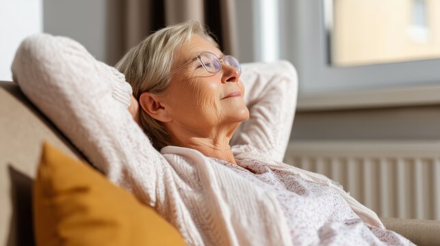 An elegant senior woman with a contemplative expression sits at home, bathed in the soft glow of natural light. She's resting on a cozy sofa, lost in thought.