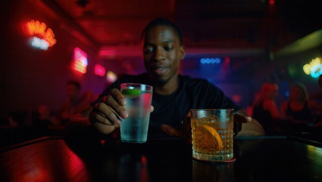 A young man sits at a bar holding a glass of water and a whiskey drink. The atmosphere is vibrant with neon lights and a lively crowd in the background.