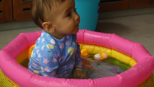 A 9 month old baby in an inflatable kiddie pool. She splashes around in the water while also being spoon fed some food.
