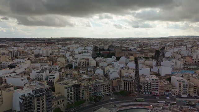 Drone view over central Malta showing the urban landscape of Ħamrun, Santa Venera, and Marsa, with dense residential buildings under dramatic cloudy skies and soft sunlight.