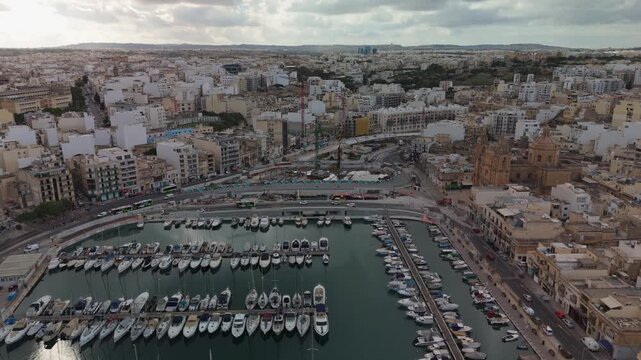Drone flight moving forward above Msida Creek, Malta, revealing the marina, St. Joseph Church, and the new flyover construction under cloudy skies in an evolving urban landscape.