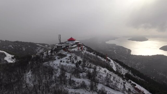 The drone pulls away from Cerro Otto&rsquo;s snowy summit station, revealing panoramic mountain and lake views above Bariloche, with Patagonian forests and clouds over Nahuel Huapi Lake.