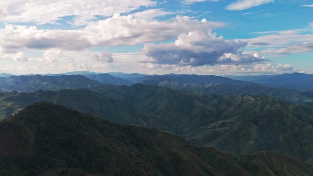 drone view over vast, layered green mountain ranges receding into a blue haze. Captured on a bright day under a beautiful blue sky with large, white cumulus clouds.Majestic and high-resolution footage