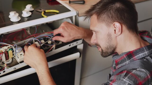 Caucasian man disconnects connector with wires using screwdriver on disassembled electric stove control panel. Portrait of focused professional repairman repairing kitchen appliance.