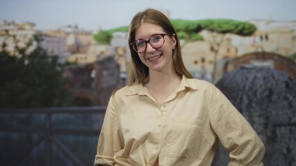Woman blonde wearing glasses smiles and waves hand at ancient roman stone ruins building outdoors; happy discovery.