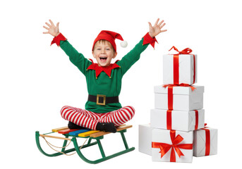 Young caucasian boy in elf costume, joyfully raising arms on colorful sled, next to white gift boxes, against transparent background, Christmas excitement and holiday celebration