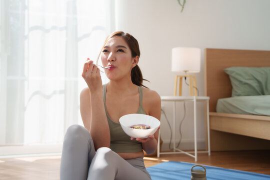 Asian woman in yoga outfit sitting on mat, happily eating fruit yogurt after workout in bright home morning light.