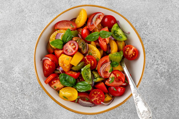 A salad of farm-fresh multi-colored tomatoes, red onions and fresh basil in a bowl on a gray concrete background. Healthy food. Top view.