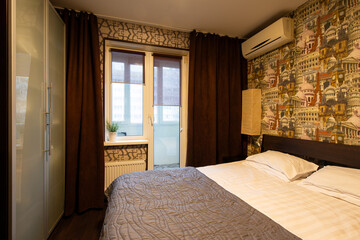Apartment interior. Double bed in the bedroom. Pillows and a bedspread on the made bed. Wallpaper with images of European landmarks. A window in the wall and a door to the balcony. Wardrobe.