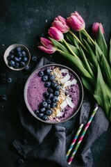 Overhead shot of a vibrant purple smoothie bowl topped with fresh blueberries and shredded coconut, accompanied by a bouquet of pink tulips and a small bowl of blueberries