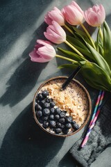 Overhead shot of a bowl of oatmeal topped with blueberries, beside a bouquet of pink tulips and colorful straws on a dark gray surface, bathed in sunlight