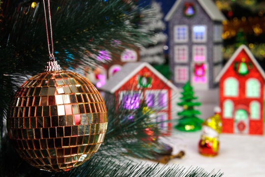 Behind the golden ball hanging on the Christmas tree branch, a Christmas village is partially blurred