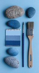 Flatlay of painted stones, paintbrushes, and a color palette on a light blue background.  The arrangement is vertical and symmetrical