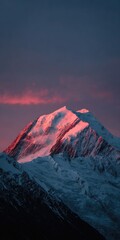 Majestic snow-capped peak illuminated by a vibrant sunset, casting a warm pink glow on its icy slopes against a dark, moody sky
