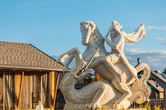 A majestic stone sculpture of St. George slaying a dragon — the knight on horseback wields a spear, poised in battle. Set against a backdrop of a wooden house and a clear blue sky, the scene combines 