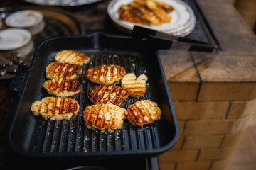 Golden-brown grilled cheese patties sizzling on a black grill pan. Visible grill marks add texture. Tongs are ready for flipping. Plates with more food in the background &mdash; perfect for food stock photo
