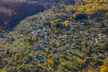 Autumn aerial of Dardhë with hillside homes, vibrant trees and layered mountain slopes lit by warm seasonal sunlight.