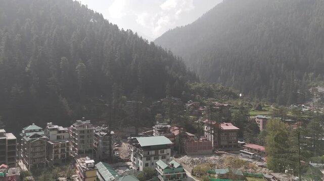 Aerial view of Kasol, Himachal Pradesh, showing village houses surrounded by autumn greenery in Parvati Valley, with vast Himalayan mountains covered in pine and deodar trees under a cloudy sky.