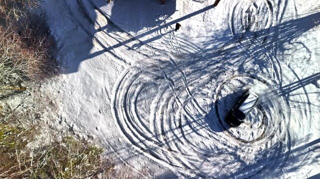 A car spins donuts in snow until it starts smoking heavily; nearby people rush to fan away the fumes in a chaotic winter mishap - top down view.