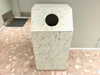 A unique, speckled white recycling bin with green and yellow flecks stands on a tiled floor, featuring a circular opening for disposal.