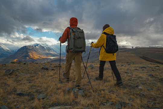 Two hikers in bright autumn gear with backpacks and trekking poles walk a mountain road overlooking a vast valley with a lake and snow-dusted peaks at golden hour.