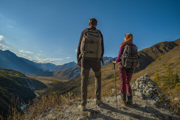 A couple of hikers with backpacks and trekking poles stand on a mountain road cliff, overlooking a vast autumn valley with mountains, forest, and a winding river below.