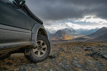 A close-up of an off-road vehicle's aggressive mud tire on a rocky ridge overlooking a valley with a lake and snow-dusted autumn mountains. Rugged tread against nature's beauty.