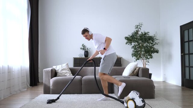 Joyful young man having fun cleaning the house, actively dancing and singing while vacuuming the carpet in his modern living room, wearing headphones and enjoying domestic chores