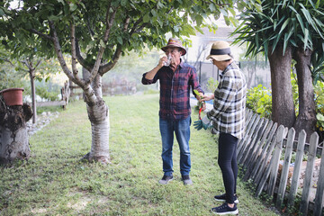 Senior couple taking a break from gardening, with the man drinking water from a glass while the woman holds a removed branch, enjoying their hobby outdoors