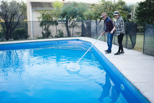 Mature couple tending their backyard pool together, skimming surface with a net on a sunny day, enjoying teamwork, maintenance and relaxed retirement lifestyle