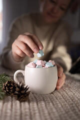 smiling woman dropping pastel marshmallow into steaming mug, knit sweater and soft focus background, pinecone