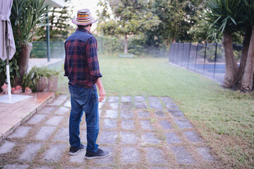 Senior man standing in his backyard garden, wearing a plaid shirt and hat, looking out over the lawn, reflecting on life and enjoying peace in his outdoor space