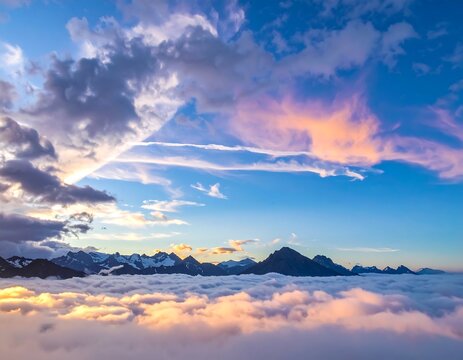 Aerial view of a mountain range emerging from a sea of clouds, illuminated by a sunset. Hues of orange, pink, and blue fill the sky