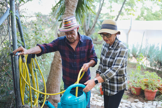 Senior couple working together in the garden, with the man operating a hose to fill a blue watering can while the woman holds it, symbolizing teamwork and healthy active retirement - Powered by Adobe